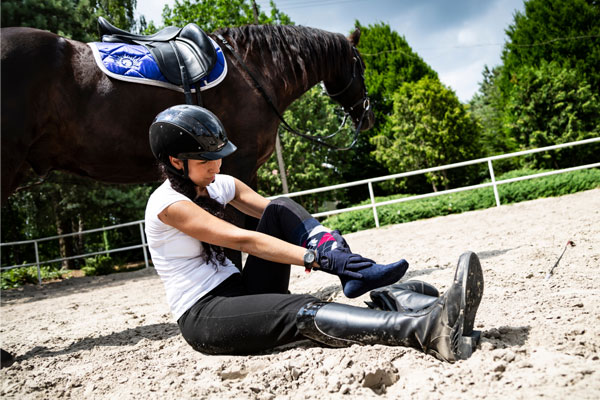 Foto einer gestürzten Reiterin auf einem Reitplatz. Sie sitzt am Boden neben ihrem Pferd und hält sich ihr linkes Bein mit den Händen fest.
