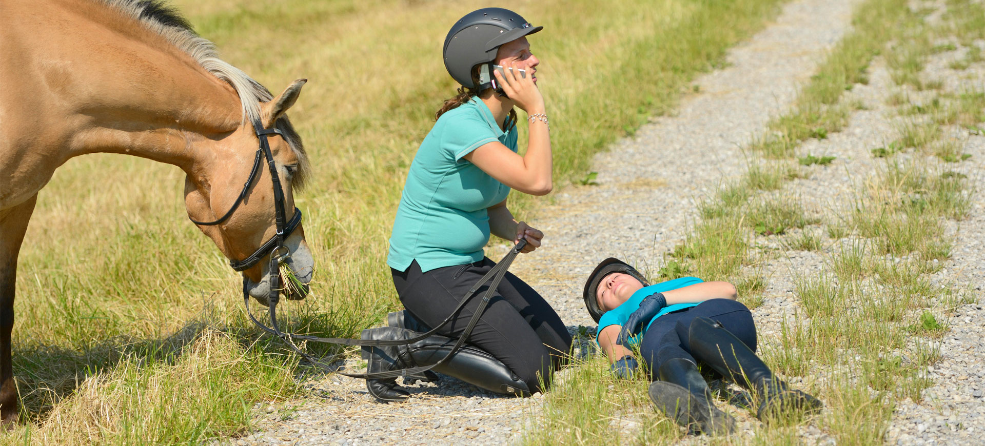 Foto eines Reitunfalls im Gelände. Neben einer bewusstlosen Reiterin kniet eine weitere Reiterin und ruft mit dem Smartphone den Rettungsdienst.